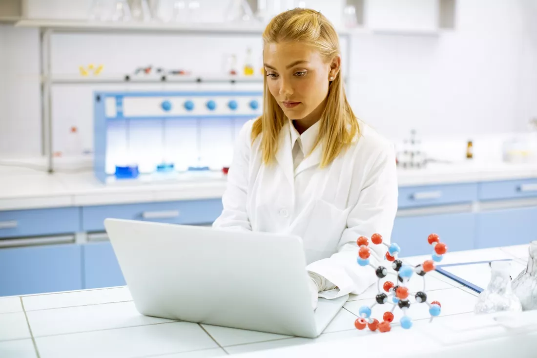 Młoda badaczka w fartuchu pracuje na laptopie obok modelu cząsteczki w laboratorium